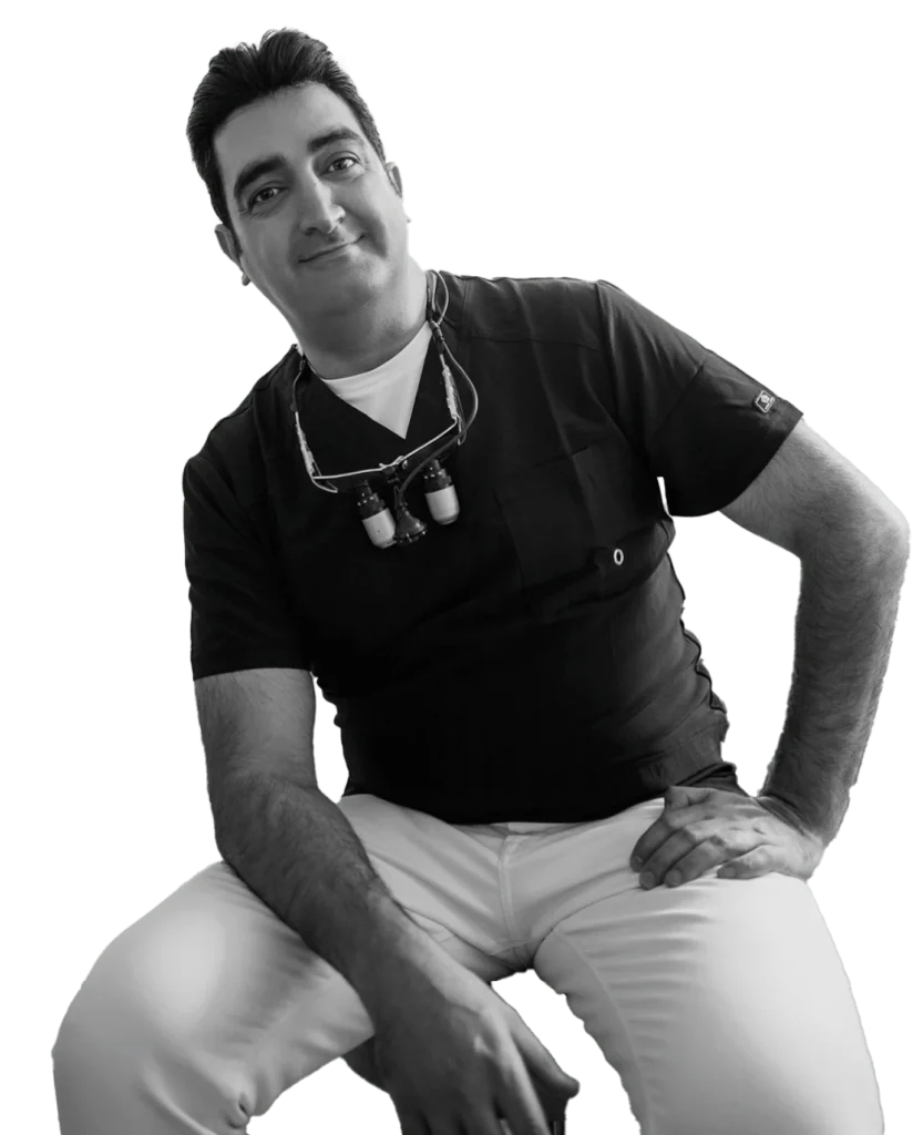 A man in black medical scrubs and white pants sits, wearing dental loupes around his neck, looking at the camera with a neutral expression.