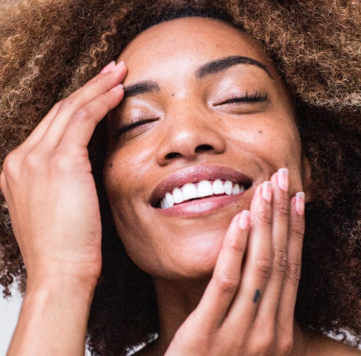 A person with curly hair smiles with closed eyes, touching their face gently with both hands.