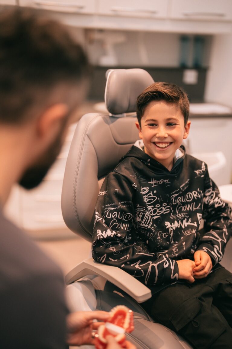 A smiling boy sits in a dental chair, facing a person holding a dental model, in a modern dental surgery.
