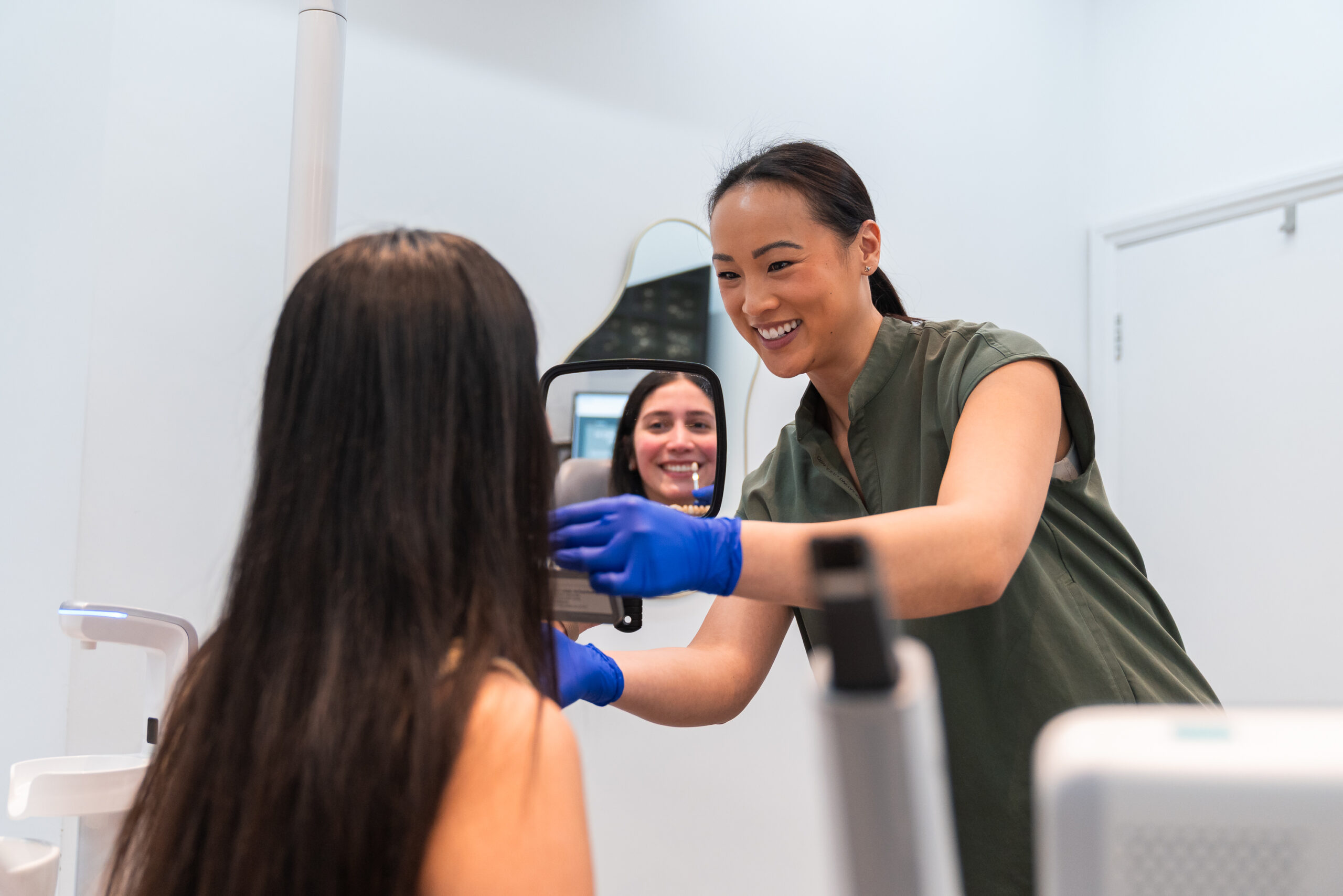 A dental professional shows a patient her reflection in a handheld mirror after teeth whitening in Hampstead, whilst another person smiles in the background.
