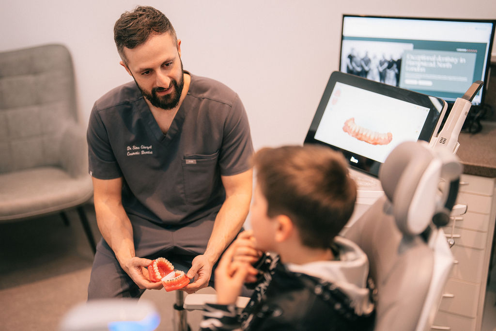 A dentist shows a child a model of teeth while sitting in a dental surgery, with dental equipment and a computer screen visible.